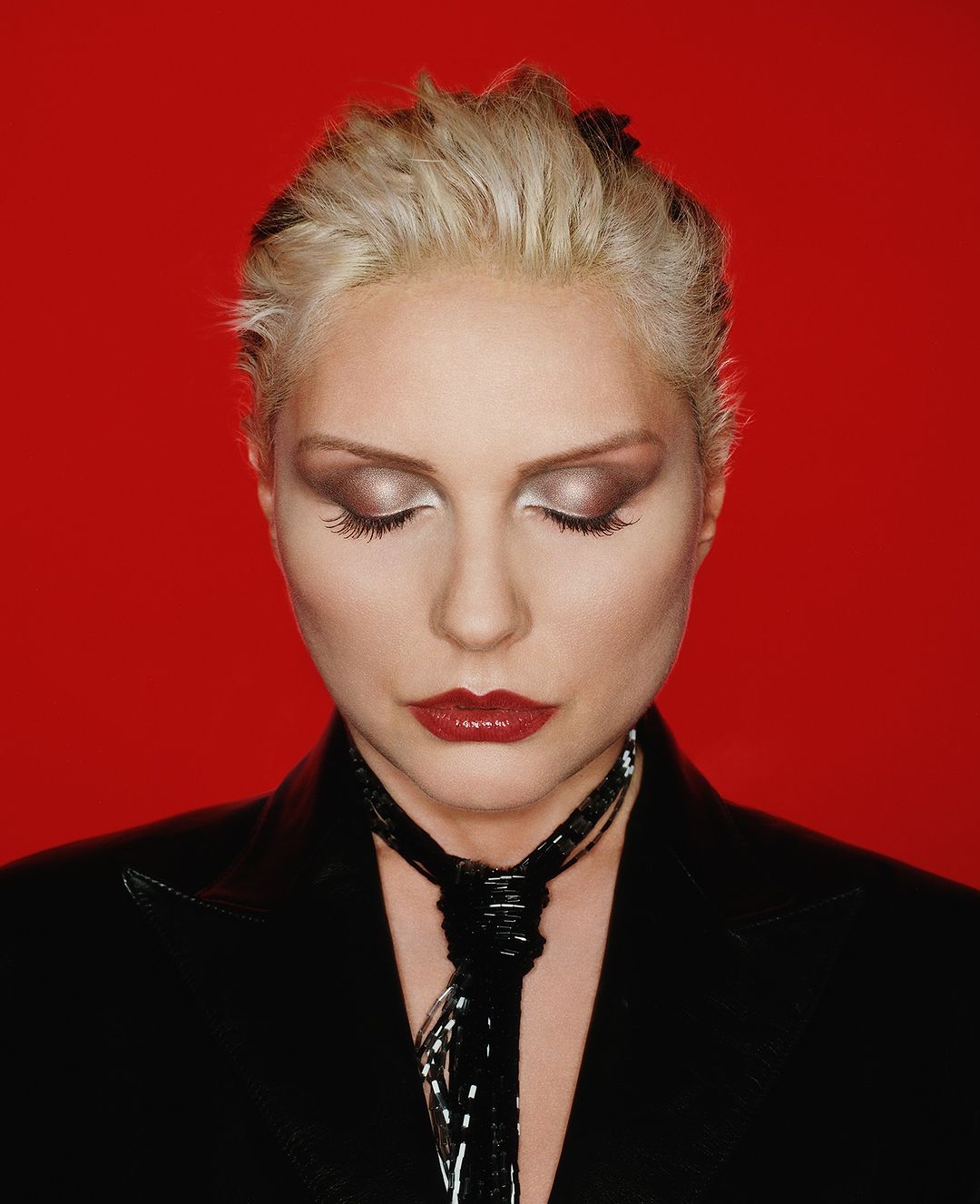Portrait of a blonde adult woman with closed eyes, wearing a black blazer and tie against a solid red background in a studio shot.