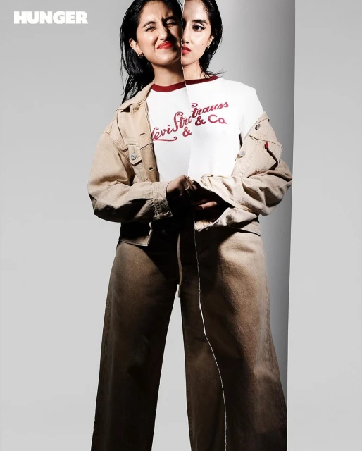 Portrait of a young woman wearing a tan jacket and pants. Part of her face is reflected against a flat mirror, with the Hunger logo.