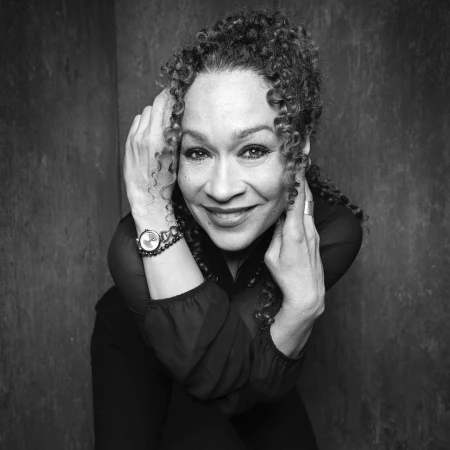Portrait of a smiling woman with curly hair wearing dark attire and a wristwatch against a textured background by Rankin.