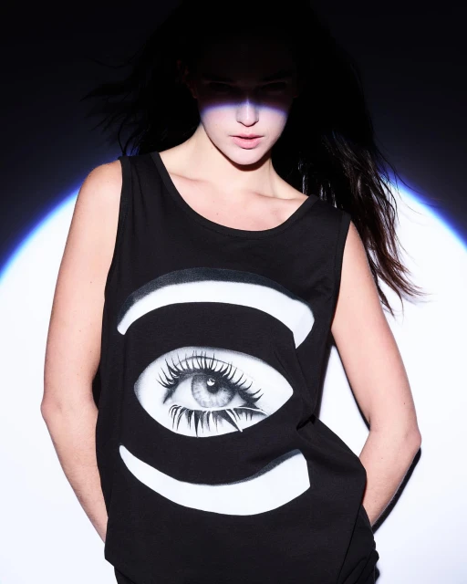 Adult woman in black tank top with eye graphic, posing in a dark, high-contrast indoor setting under a spotlight.