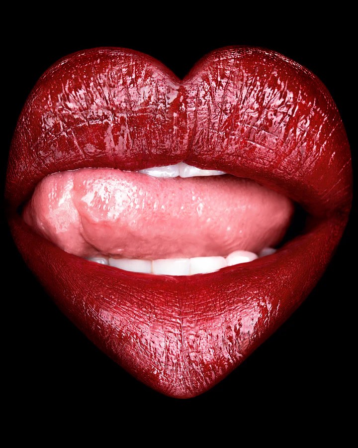 Close-up studio shot of a person's mouth with red lipstick formed like a heart shape, slightly open with the tongue visible against a black background.