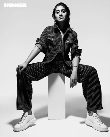 Black and white studio shot of a model wearing a striped denim jacket and pants, posing on a white cube with white sneakers.