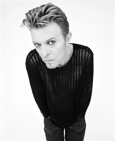 Portrait of David Bowie leaning forward wearing a black knit sweater against a white seamless background