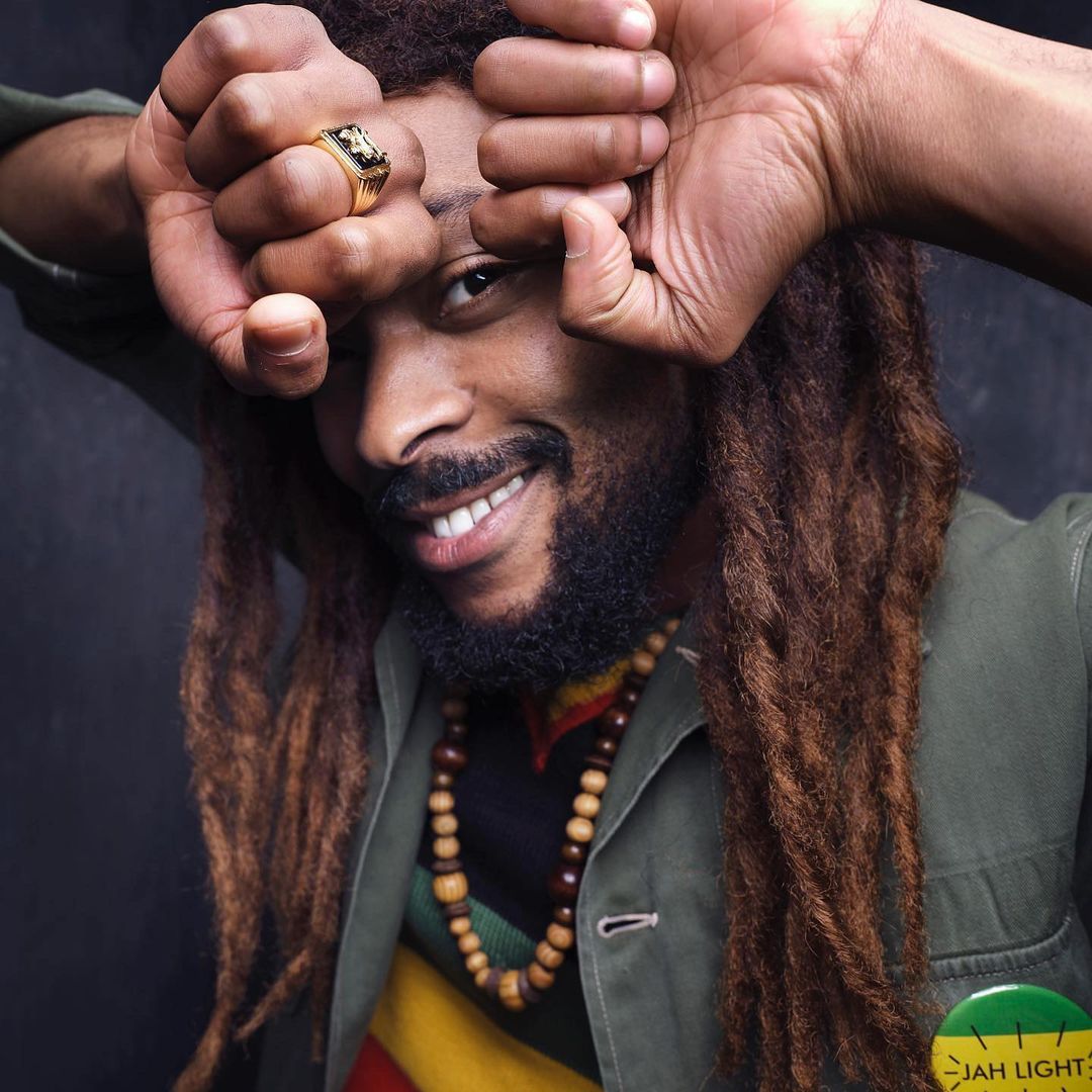 Close-up portrait of a Black man with long dreadlocks and a beard, smiling while holding both fists up near his forehead.