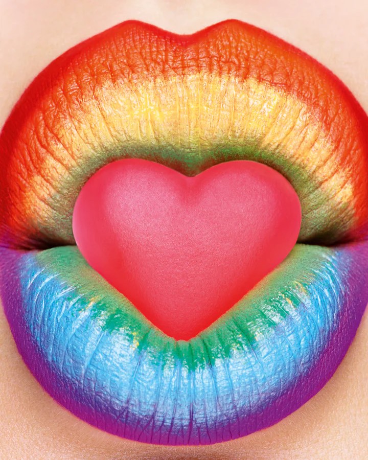 Close-up of rainbow-colored lips holding a pink heart-shaped candy, creating a vibrant and expressive beauty image.