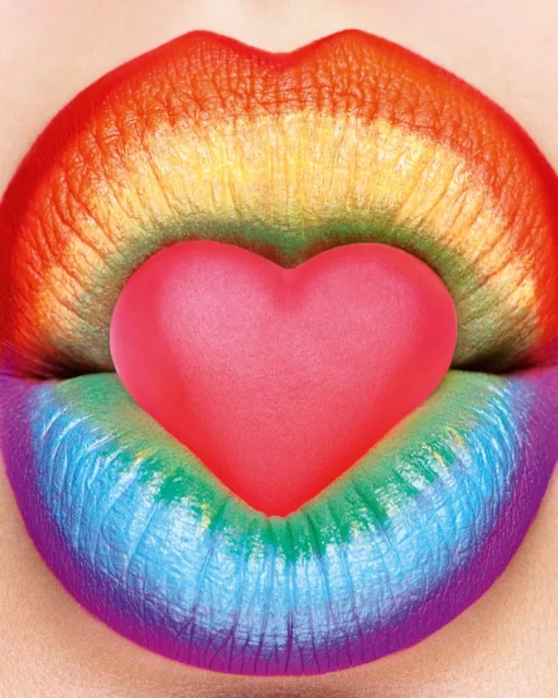 Close-up of rainbow-colored lips holding a pink heart-shaped candy, creating a vibrant and expressive beauty image.