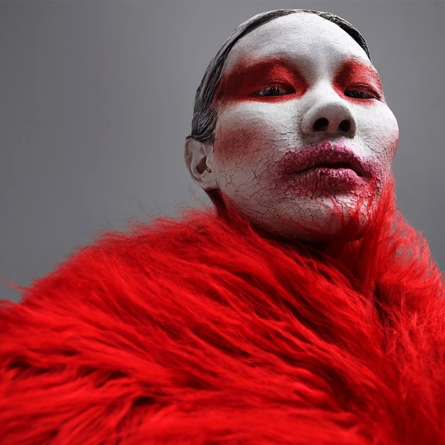 Striking close-up portrait of a person with cracked white facial makeup and red accents, wearing a bright red, shaggy faux fur stole.