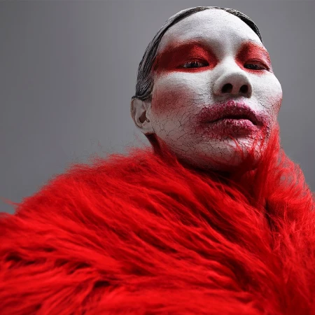 Striking close-up portrait of a person with cracked white facial makeup and red accents, wearing a bright red, shaggy faux fur stole.