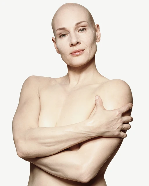 Bald adult woman poses with arms crossed in a studio portrait. Head and shoulders shot against a white background with soft studio lighting.