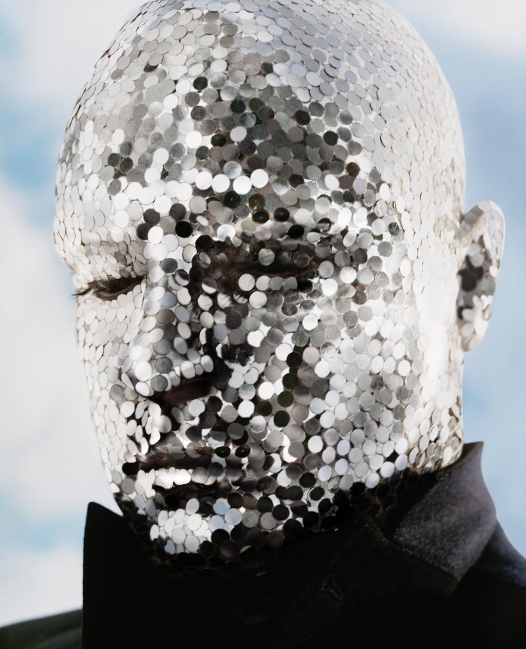 Close-up portrait of a male with silver disc makeup, wearing a black suit jacket and bow tie against a blurred sky background.