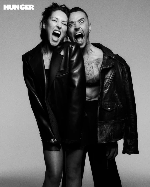 Black and white portrait of a woman in a blazer and a man in a leather jacket yelling with mouths open against a neutral backdrop.