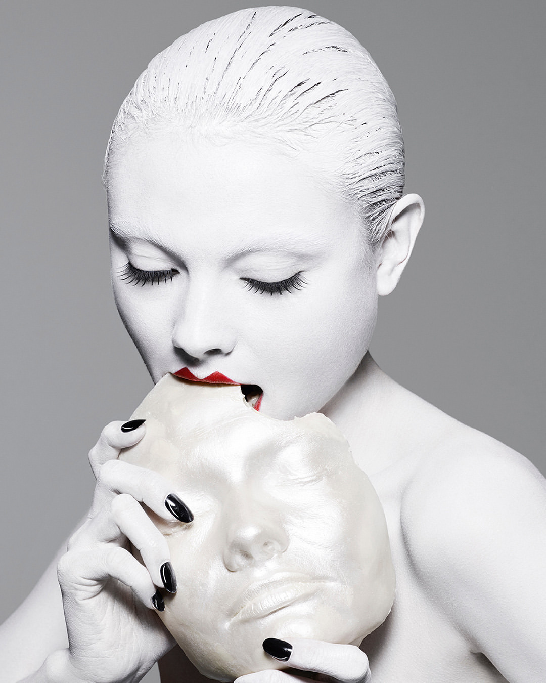 A woman with white face paint and red lipstick holds a white mask to her face in a studio shot against a gray background.