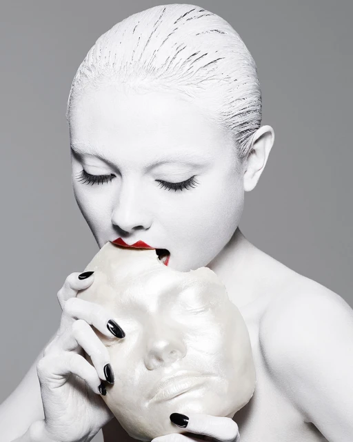 A woman with white face paint and red lipstick holds a white mask to her face in a studio shot against a gray background.