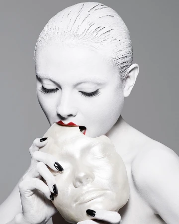 A woman with white face paint and red lipstick holds a white mask to her face in a studio shot against a gray background.