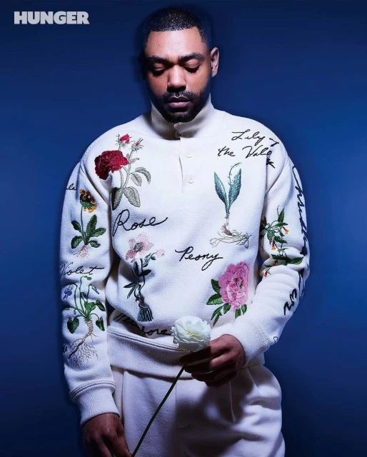 Portrait of a man in an embroidered sweater holding a white flower against a dark blue background in a studio setting.