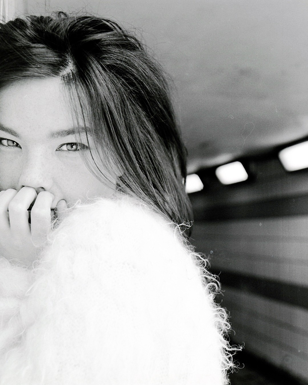 Monochrome portrait of a woman indoors with shoulder-length hair, wearing a fluffy jacket, covering her mouth with her hand in a tunnel.
