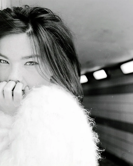 Monochrome portrait of a woman indoors with shoulder-length hair, wearing a fluffy jacket, covering her mouth with her hand in a tunnel.
