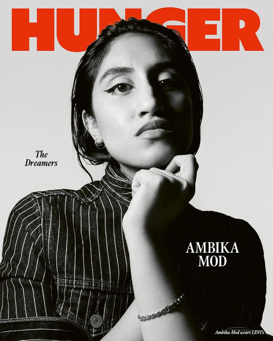 Close-up studio shot of Ambika Mod in a striped top, Hunger Magazine cover, neutral backdrop. The cover features the text 'HUNGER' and 'AMBICA MOD'.