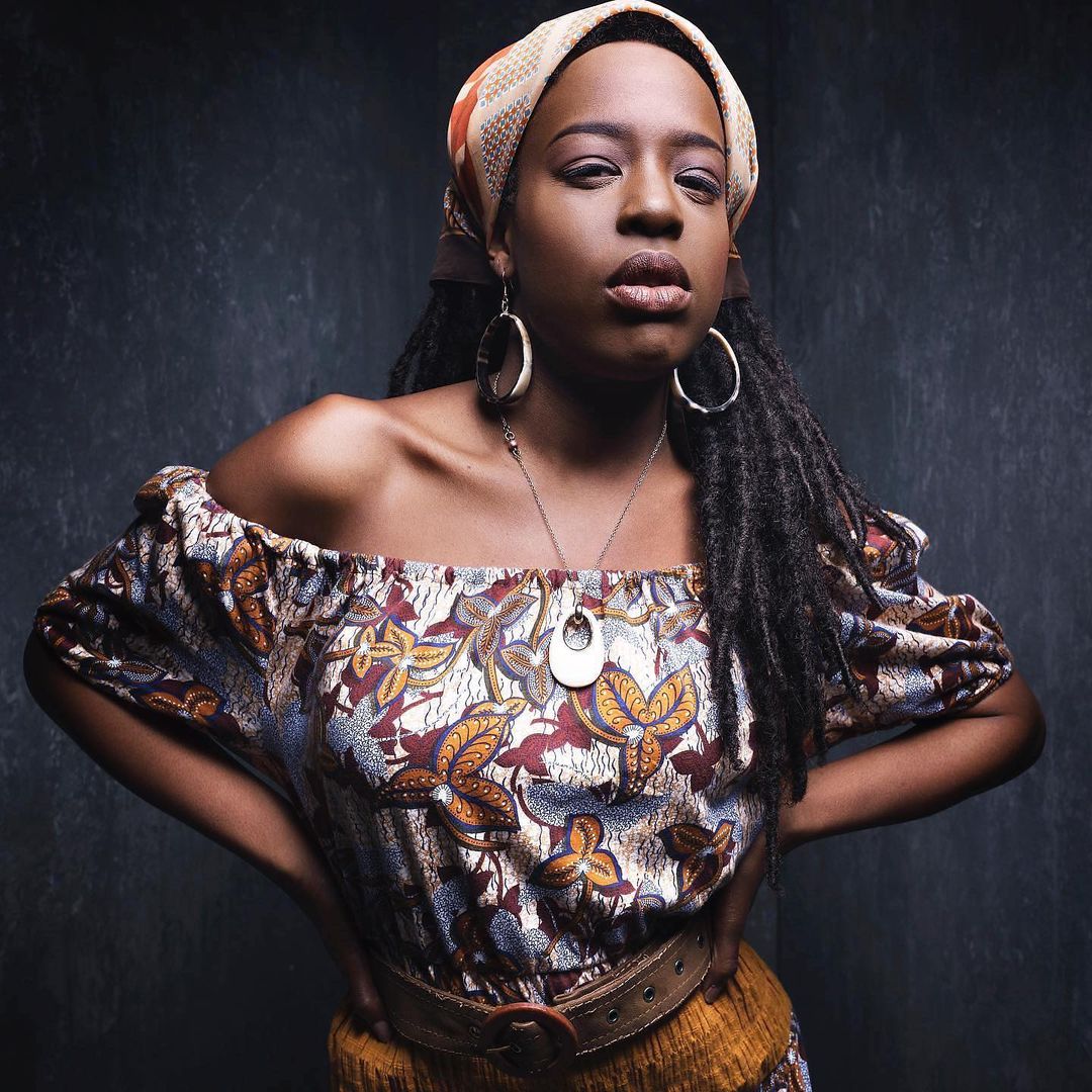 Portrait of an African-American woman wearing an off-the-shoulder floral print top, a tan headscarf, and large hoop earrings, against a dark background.