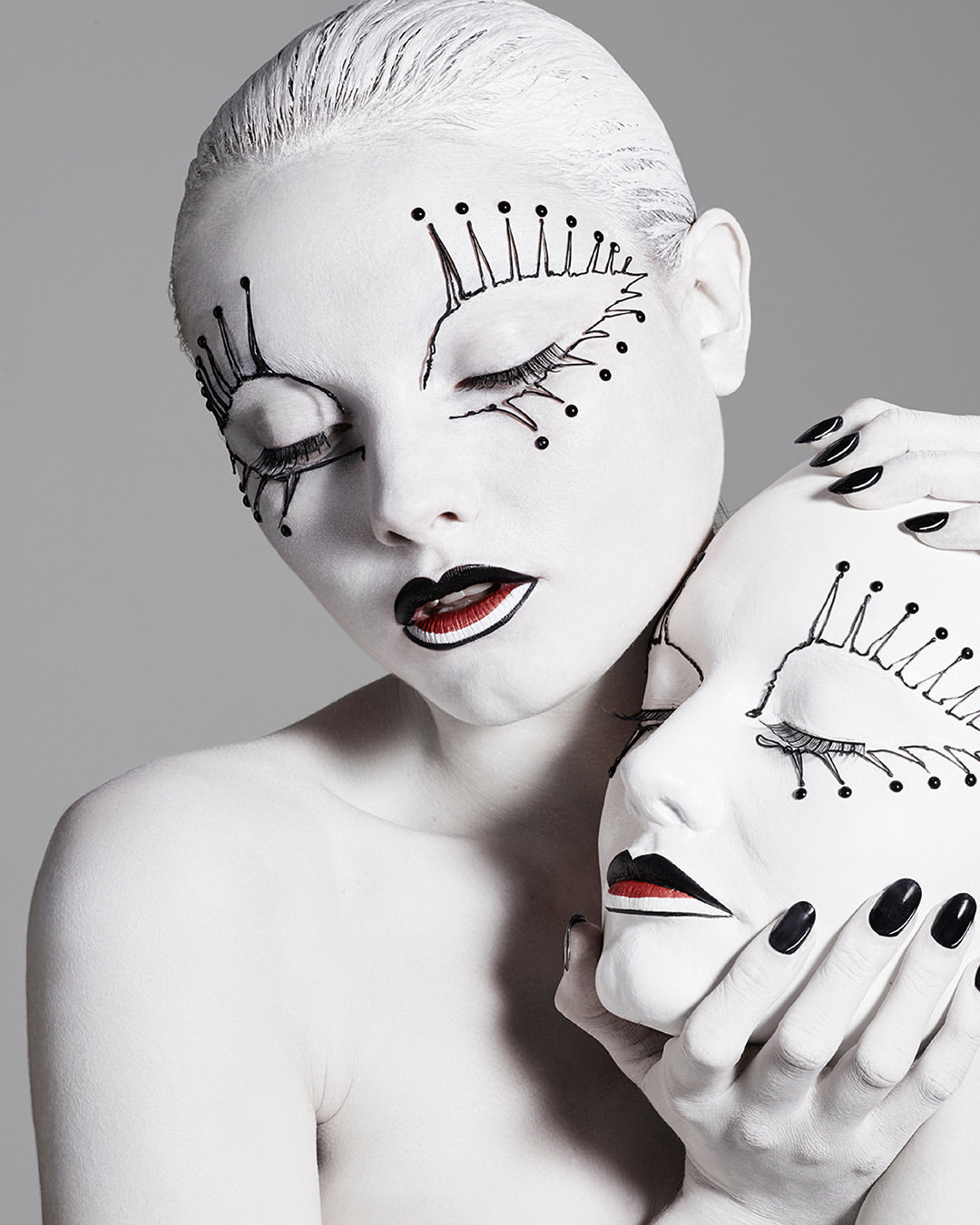 Artistic portrait of a model with painted white skin, black lips, and theatrical makeup, holding a matching white mask.
