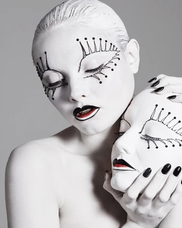 Artistic portrait of a model with painted white skin, black lips, and theatrical makeup, holding a matching white mask.