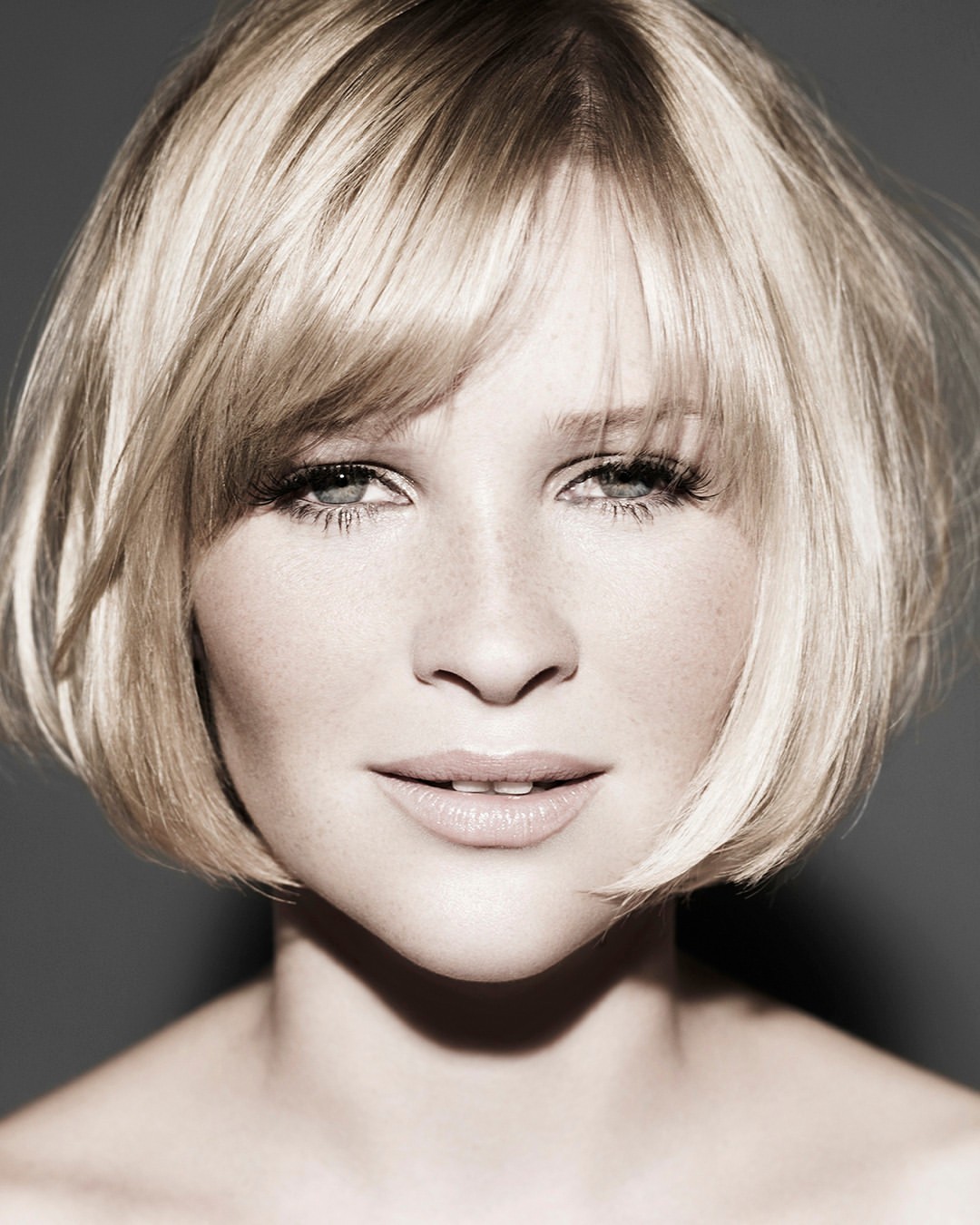 Close-up portrait of a woman with short blonde bob hair and bangs, looking directly at the camera against a neutral dark grey background.