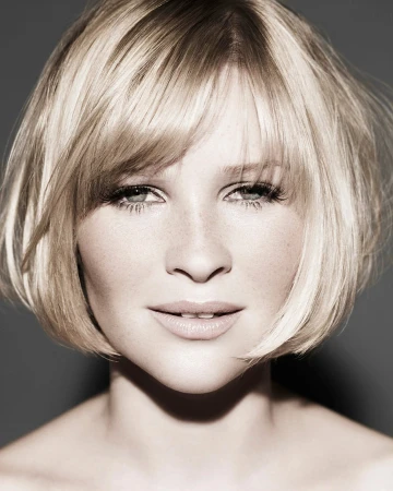 Close-up portrait of a woman with short blonde bob hair and bangs, looking directly at the camera against a neutral dark grey background.