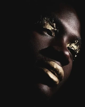 Close-up portrait of a woman with dark skin, gold foil makeup on her eyes and lips, and dramatic lighting against a dark background.
