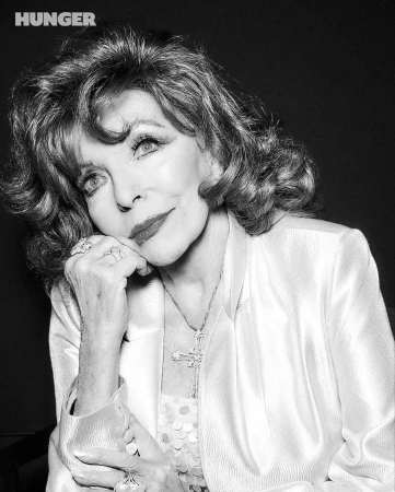 Monochrome portrait of Joan Collins resting her chin on her hand, wearing a light satin blazer and crucifix pendant.