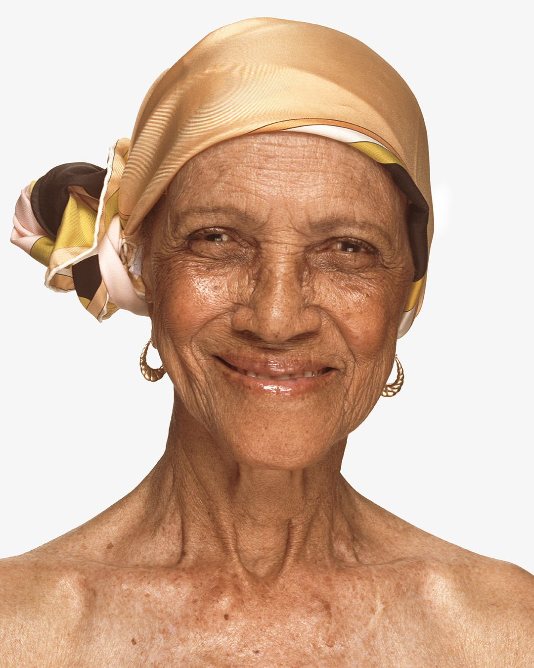 Close-up portrait of a smiling elderly woman with tan skin, wearing gold hoop earrings and a patterned silk headscarf against white.