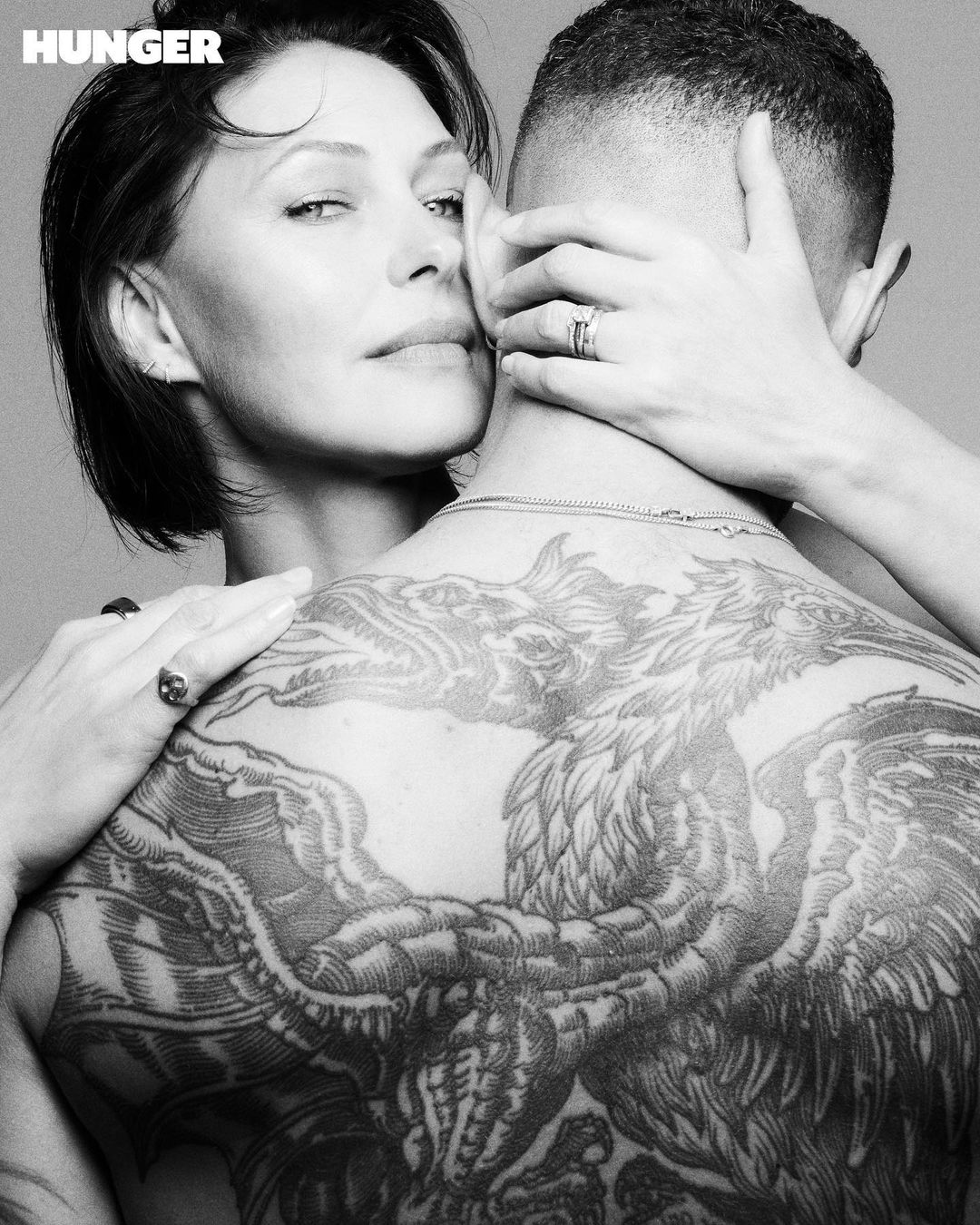 Black and white portrait of a woman embracing a tattooed man with a back tattoo, Hunger magazine logo visible in the upper left corner.