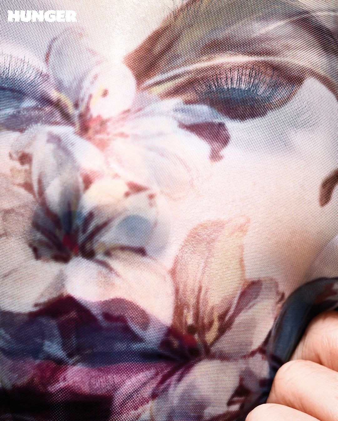 Close-up studio shot of a human face partially covered by a floral-patterned translucent fabric, highlighting an eye and fingers.