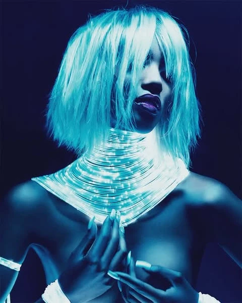 Woman with an afro and glowing light tube necklace poses against a dark background in a beauty shot with blue color grading.