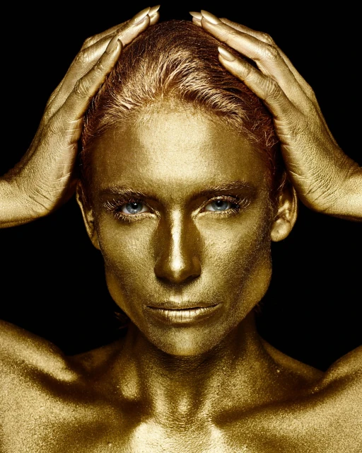Golden-skinned woman with hands on head in close-up studio portrait against black backdrop, creating a striking, high-contrast visual.