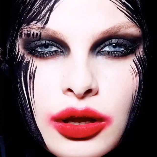 Close-up of a woman's face with dramatic black and red makeup, including exaggerated eyeliner and bold red lipstick, against a dark background.