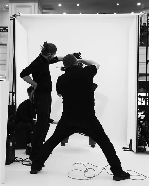 Three adults in black clothing work around a camera and white backdrop in an indoor photo studio setting during a photography session.