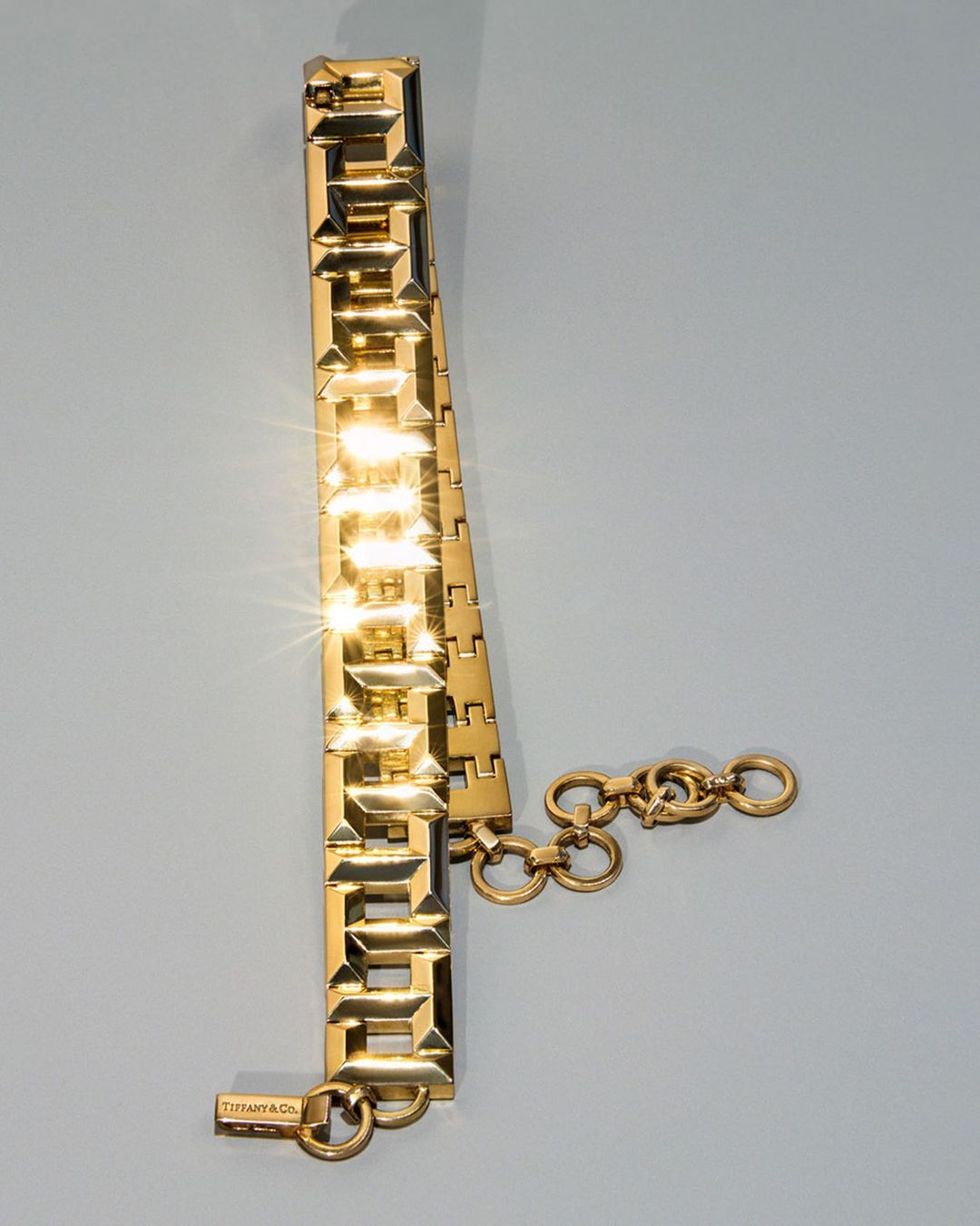 Close-up studio shot of a gold Tiffany & Co. link bracelet featuring rectangular geometric designs and a clasp on a pale blue background.