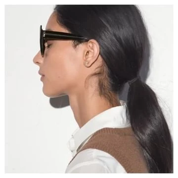 Profile portrait of a young woman with dark hair in a ponytail wearing black sunglasses and a tan vest against a white wall.