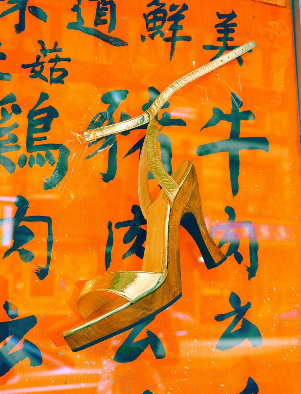 Gold high-heeled sandal displayed against an orange wall with black Asian characters, creating a fusion of fashion and culture.