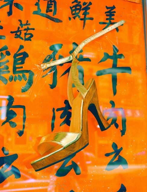 Gold high-heeled sandal displayed against an orange wall with black Asian characters, creating a fusion of fashion and culture.