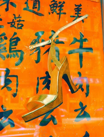 Gold high-heeled sandal displayed against an orange wall with black Asian characters, creating a fusion of fashion and culture.