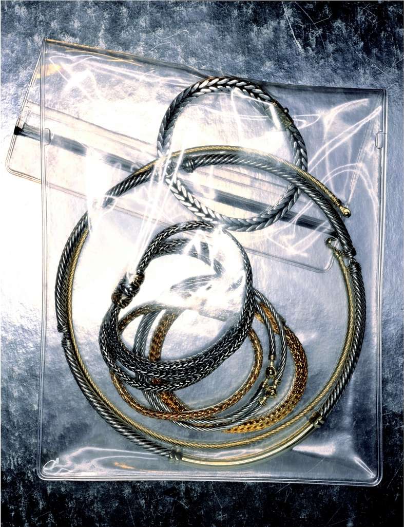 A variety of necklaces and bracelets in silver and gold tones are arranged inside a clear plastic bag, creating a textured, metallic still life.