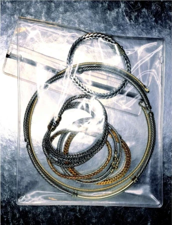 A variety of necklaces and bracelets in silver and gold tones are arranged inside a clear plastic bag, creating a textured, metallic still life.