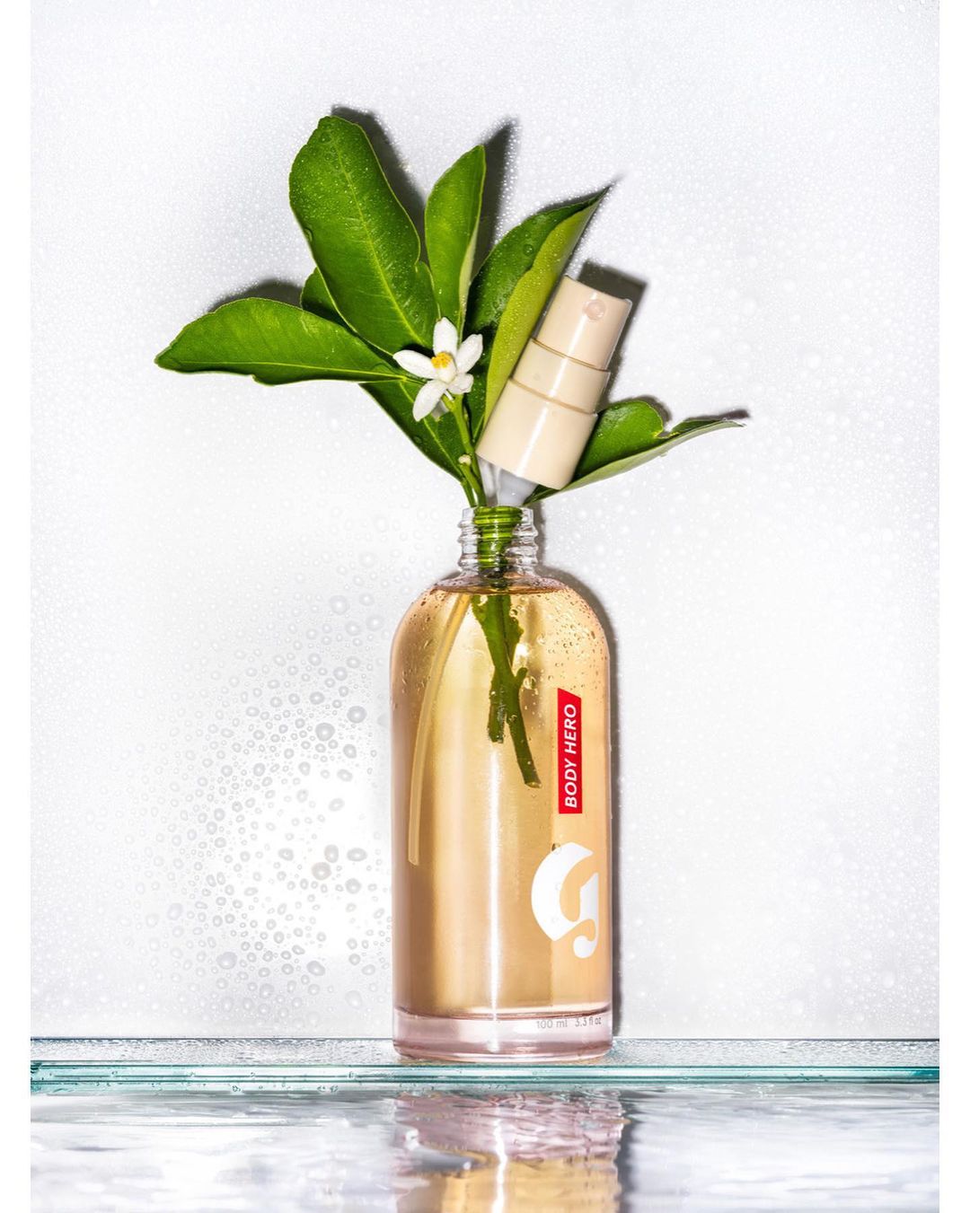 Body care product bottle with liquid inside, a white flower, and green leaves against a white background with water droplets. 