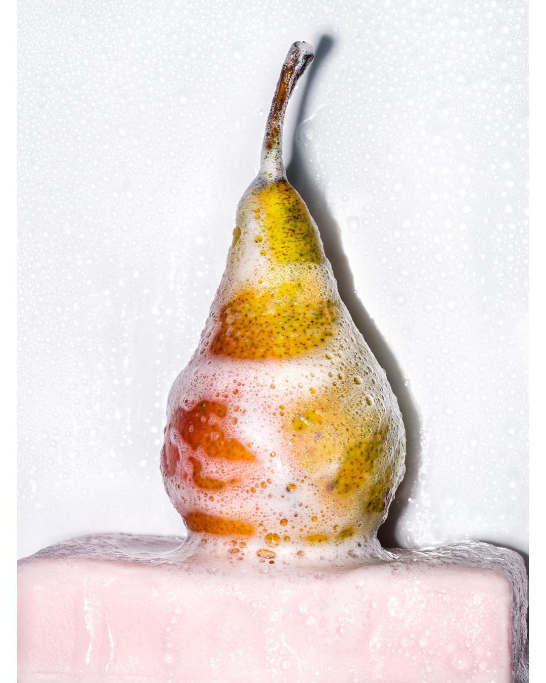 A single pear, partially covered in soap or foam, rests on a pink block against a bright white backdrop, creating a clean and minimalist still life.