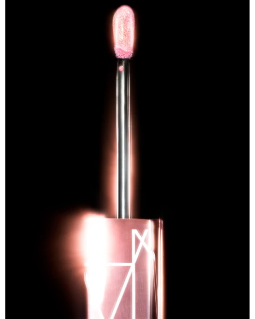 Close-up of a lip gloss applicator with a pale pink, shimmery product on the tip, set against a dark black background.