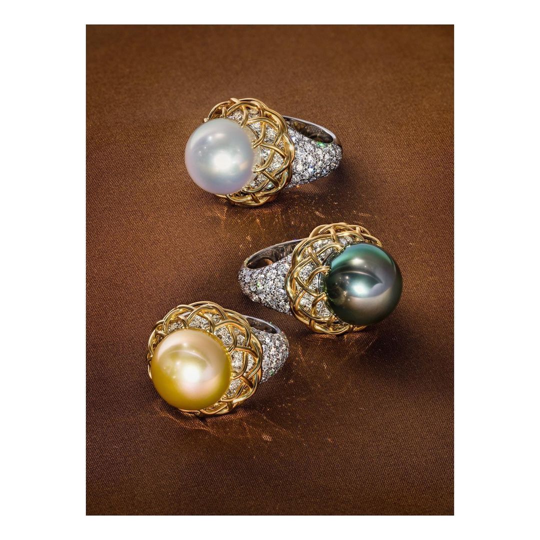 Three pearl rings with diamond-encrusted bands on a brown surface. The rings have white, black, and gold pearls.