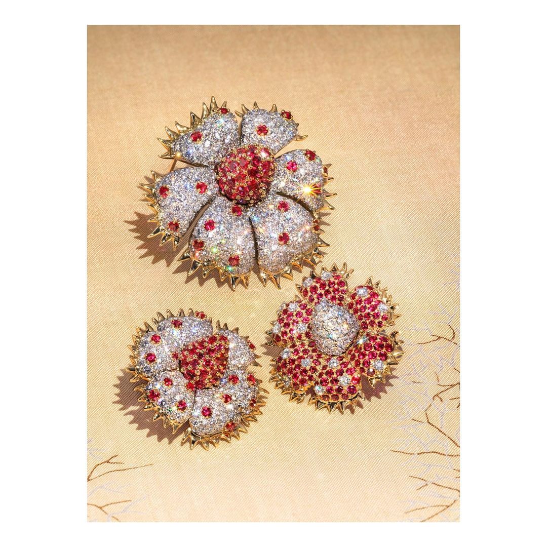 Three flower-shaped brooches sparkle with white and red stones on a textured gold background, catching the light with glittering facets.