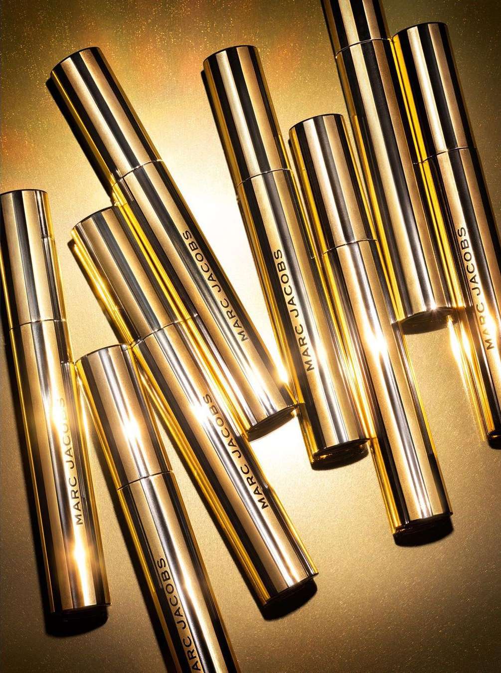 Several gold tubes of Marc Jacobs beauty product are grouped together on a gold background, product shot, luxury beauty product