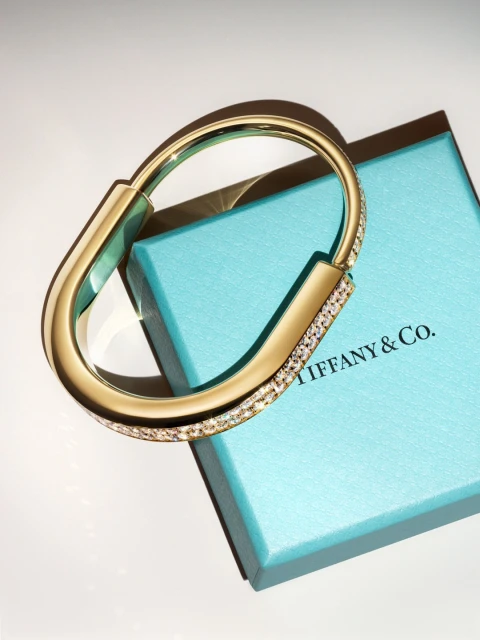 Gold bangle bracelet featuring encrusted diamonds placed on top of a Tiffany & Co. blue box against a neutral colored backdrop.
